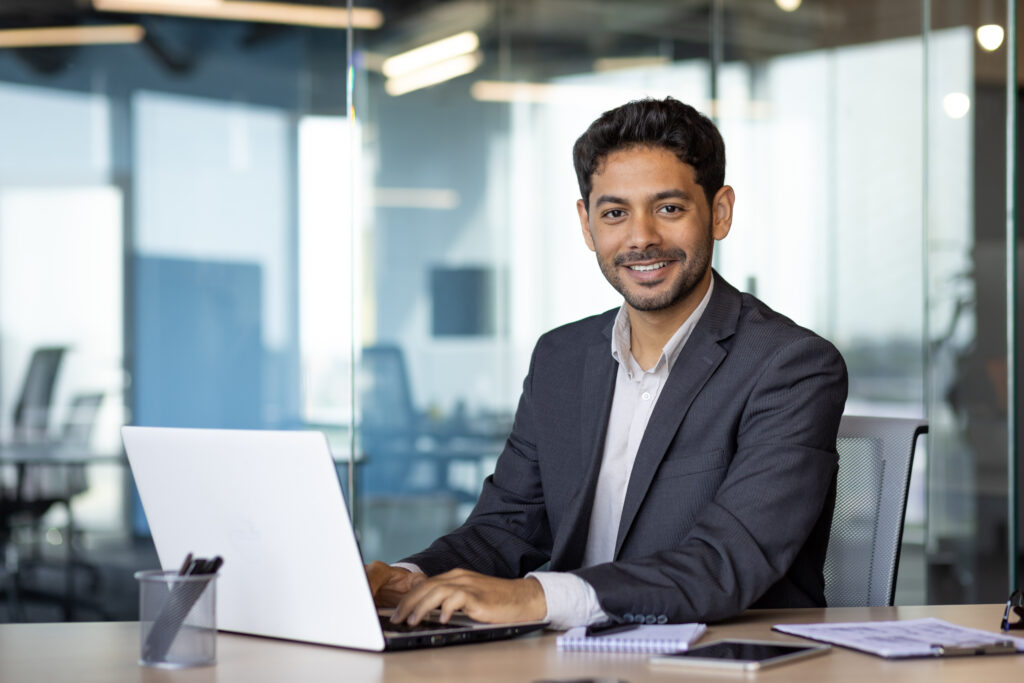 Portrait d'un jeune homme d'affaires arabe, homme souriant et regardant la caméra assis à l'intérieur d'un bureau, patron en costume d'affaires sur son lieu de travail utilisant un ordinateur portable.