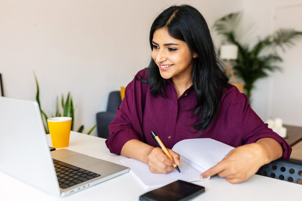 Jeune femme indienne étudiante prenant des notes en utilisant un ordinateur portable à la maison. Femme millénaire d'origine ethnique apprenant en ligne en écoutant un appel vidéo virtuel. Concept d'affaires et d'éducation.